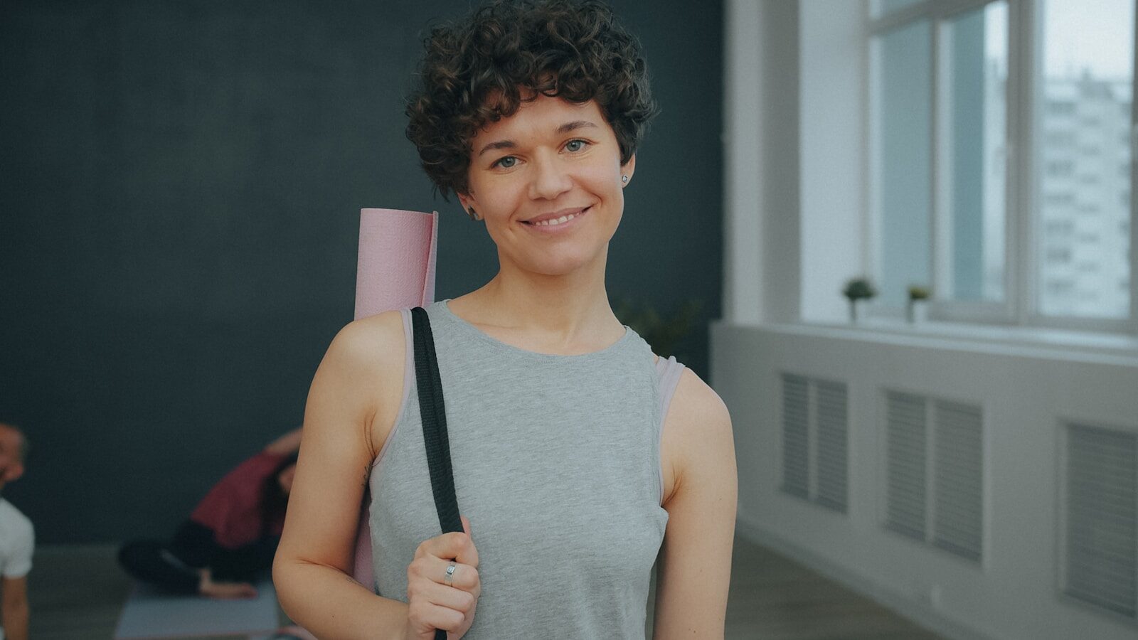 A woman with short curly hair carrying a pink yoga mat in a studio